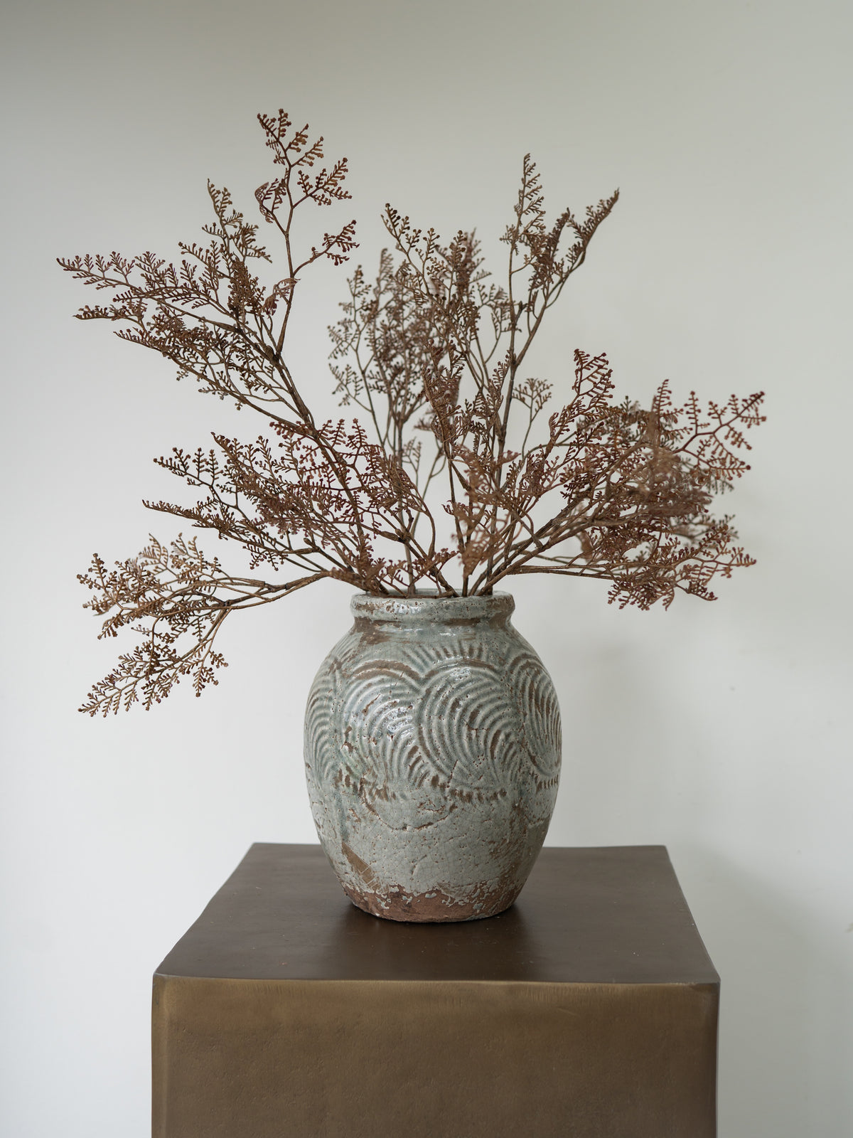 A textured ceramic vase with embossed concentric wave patterns, filled with dried brown botanical stems, displayed on a dark brown pedestal against a plain off-white wall.