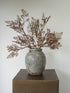 A textured ceramic vase with embossed concentric wave patterns, filled with dried brown botanical stems, displayed on a dark brown pedestal against a plain off-white wall.