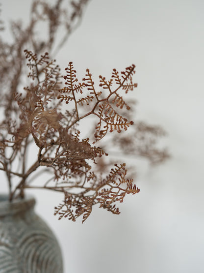 Dried brown botanical stems arranged in a textured light blue-gray ceramic vase against a soft, out-of-focus light gray background.