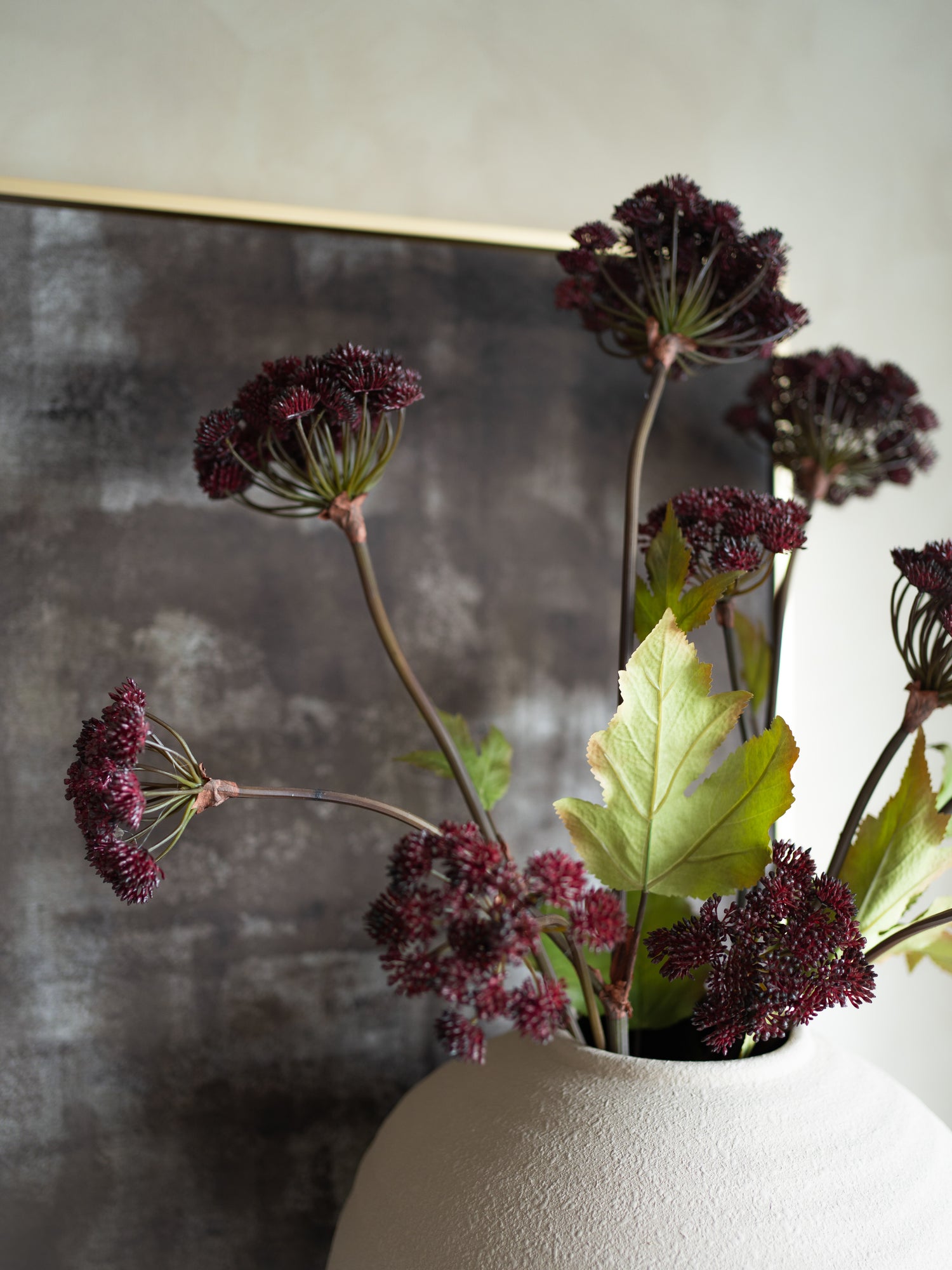 Artificial plants with dark purple flowers and green leaves in a white vase against a textured wall.