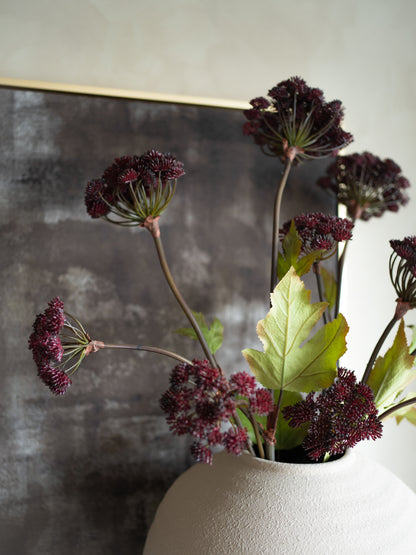 Artificial plants with dark purple flowers and green leaves in a white vase against a textured wall.