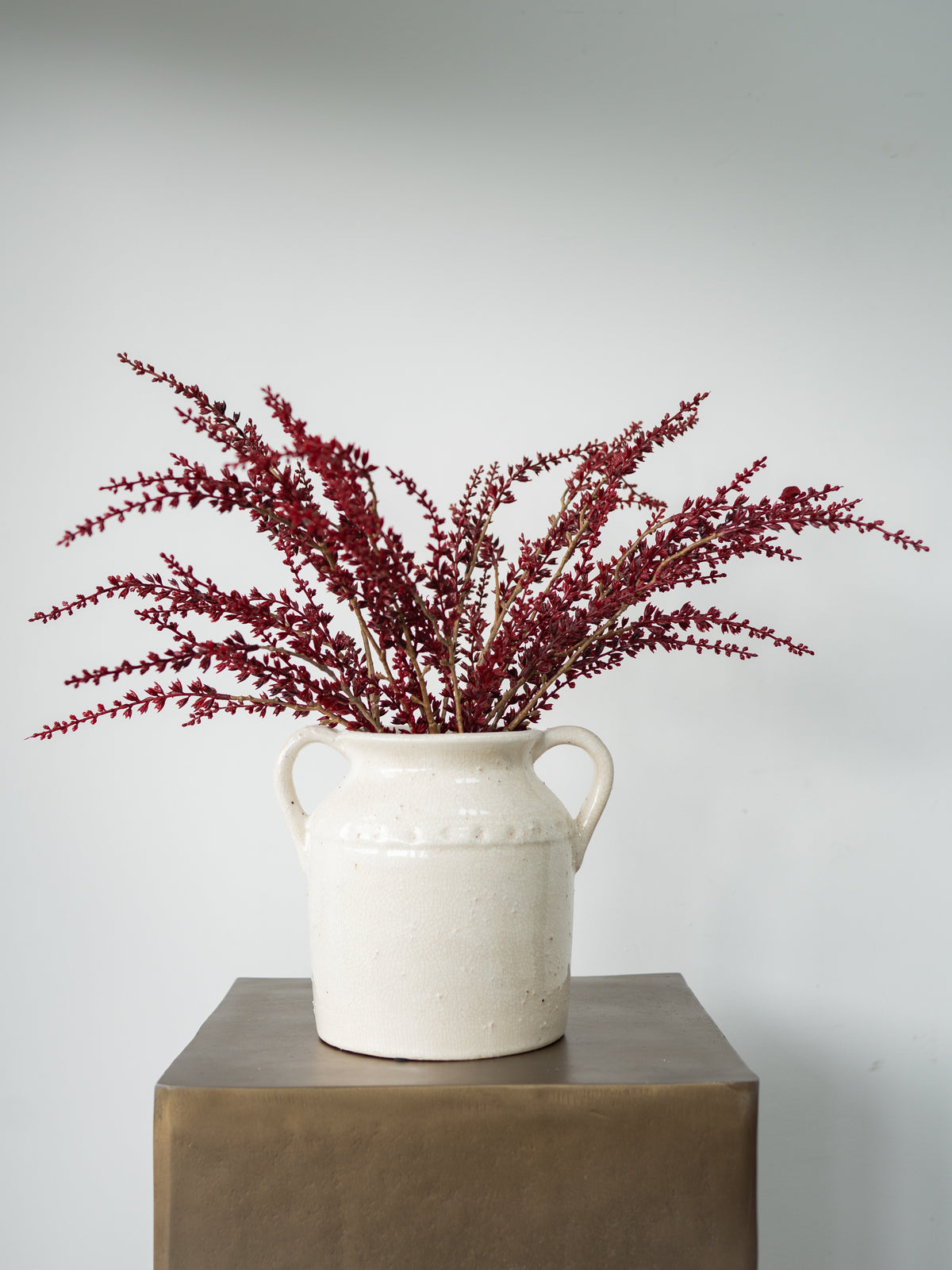 A white ceramic milk jug vase filled with dark burgundy dried heather stems, displayed on a square bronze pedestal against a light gray background.
