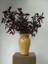 A dark burgundy leafy stem arrangement in a glossy mustard-yellow ceramic vase, displayed on a square metallic pedestal against a plain light gray wall.