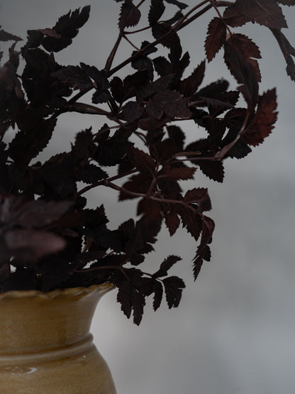 Dark burgundy artificial foliage with serrated leaves arranged in a light beige ceramic vase against a soft gray background.