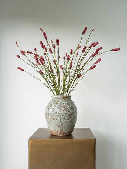 A ceramic vase with a textured, patterned glaze in muted gray-green tones, filled with dried pinkish-red flower stems, displayed on a square bronze pedestal against a light gray background.