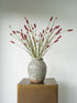 A ceramic vase with a textured, patterned glaze in muted gray-green tones, filled with dried pinkish-red flower stems, displayed on a square bronze pedestal against a light gray background.