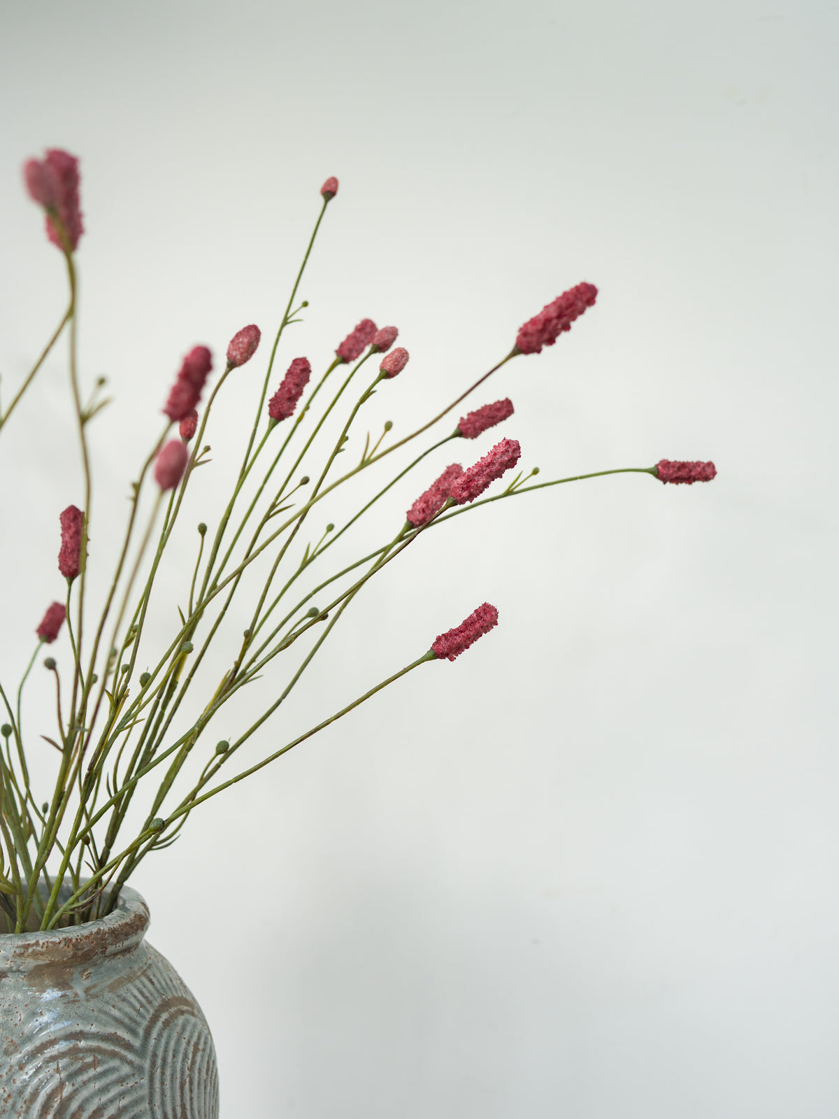 Dried pink flowers with long green stems arranged in a textured gray ceramic vase against a plain light gray background.