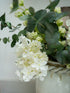 Faux white butterfly hydrangea and green leaves in a woven basket against a light background