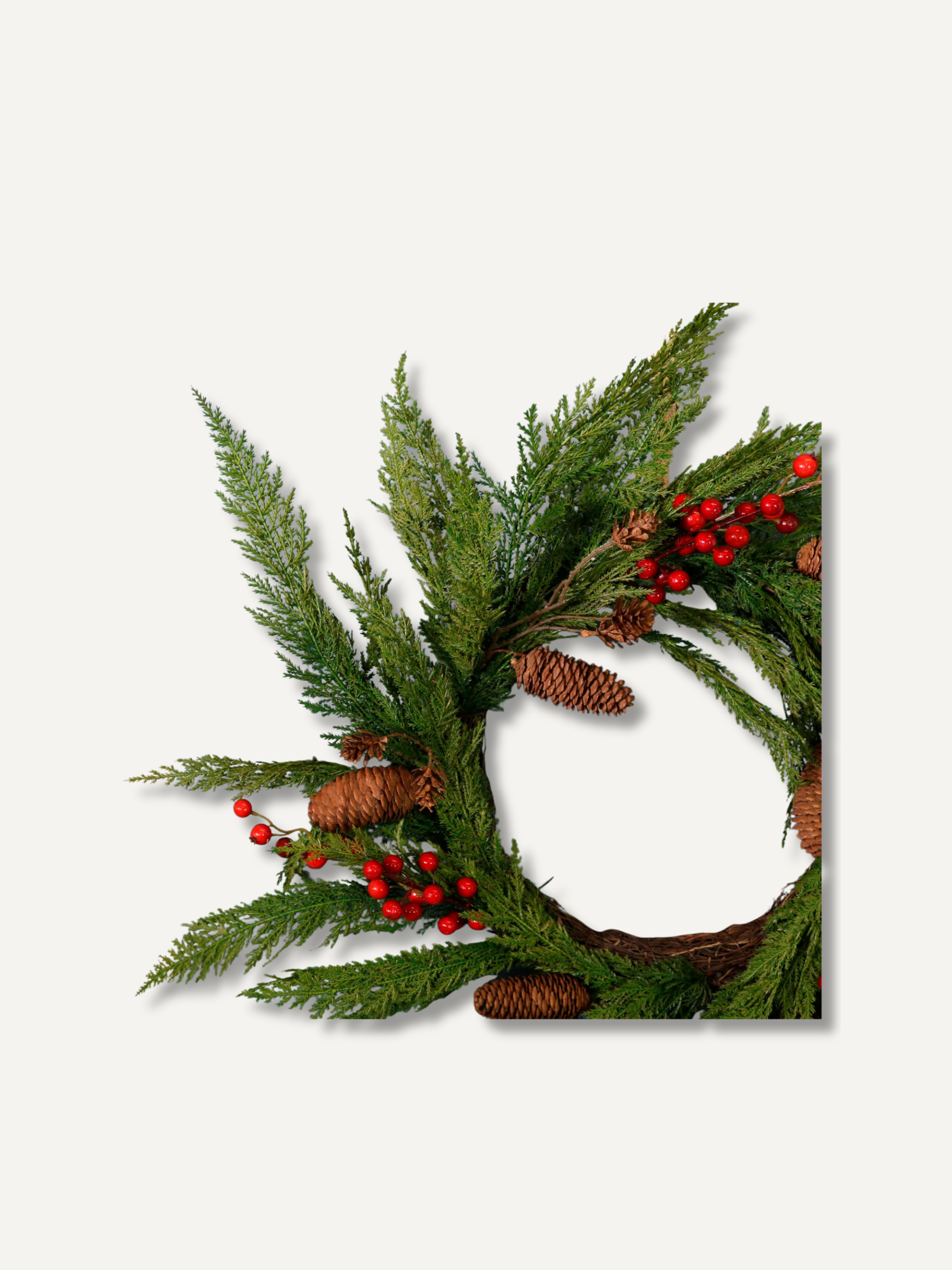 A artficial Christmas wreath made of green pine and cypress branches, decorated with small brown pinecones and clusters of red berries, shown against a plain light gray background.