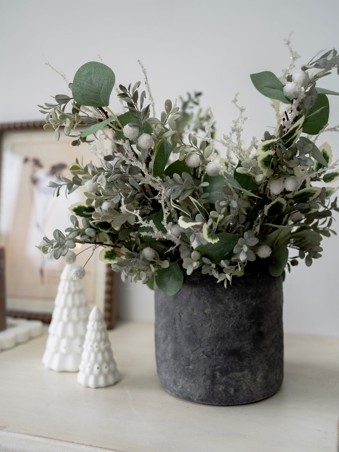 Decorative arrangement of faux christmas greenery in a gray vase on a white surface with a blurred background.