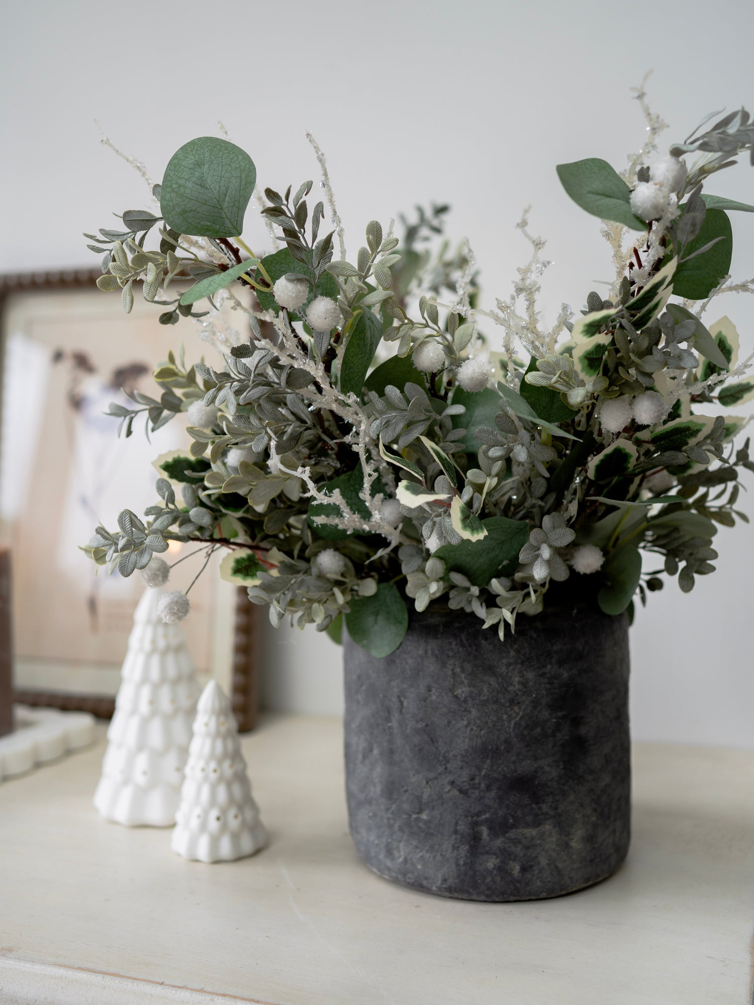 Decorative arrangement of faux christmas greenery in a gray vase on a white surface with a blurred background.
