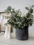 Decorative arrangement of faux christmas greenery in a gray vase on a white surface with a blurred background.