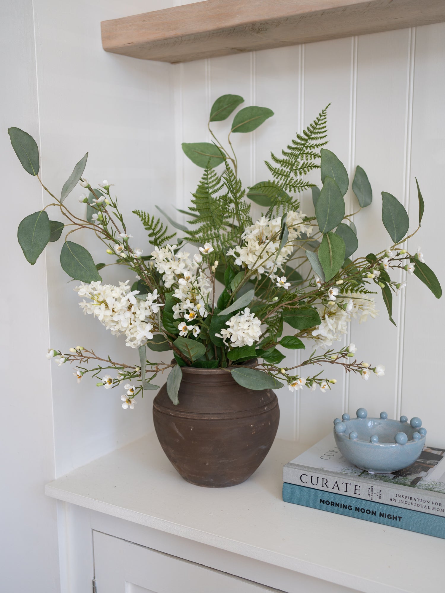 Decorative arrangement of faux white flowers and greenery and white flowers in a brown vase on a shelf.