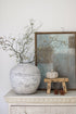 Decorative setting with a textured vase, branches, a pumpkin, and a small stool on a white surface.