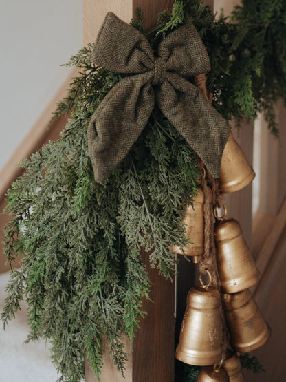 Decorative faux cypress garland with gold bells and a brown bow on a wooden surface.