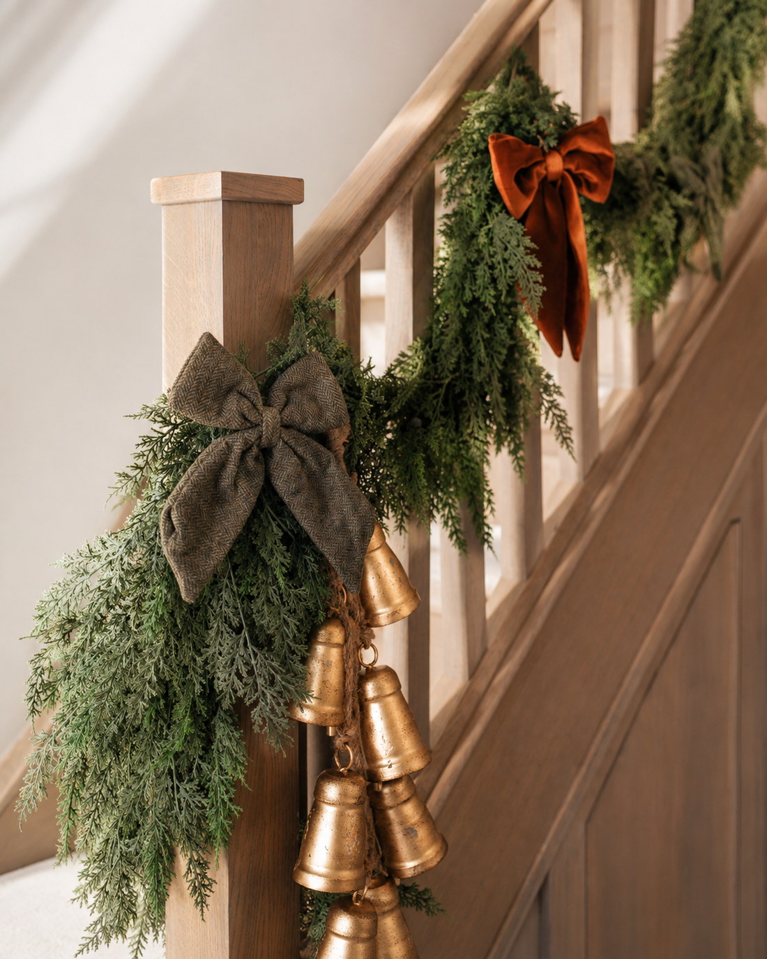 A christmas stair rail decorated with green garlands, large fabric bows in dark taupe and rust orange, and a cluster of metallic gold bells.