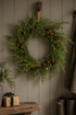 A faux green pinecone garland wreath hanging on a dark gray paneled wall, with a rustic wooden shelf and wrapped gift box below.