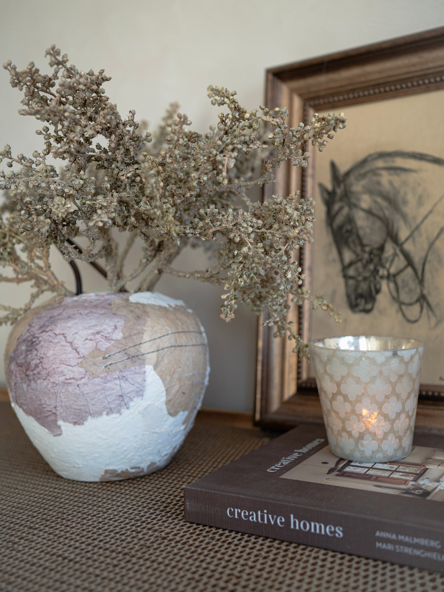 Decorative vase with dried flowers on a surface with a book and candle in the background.