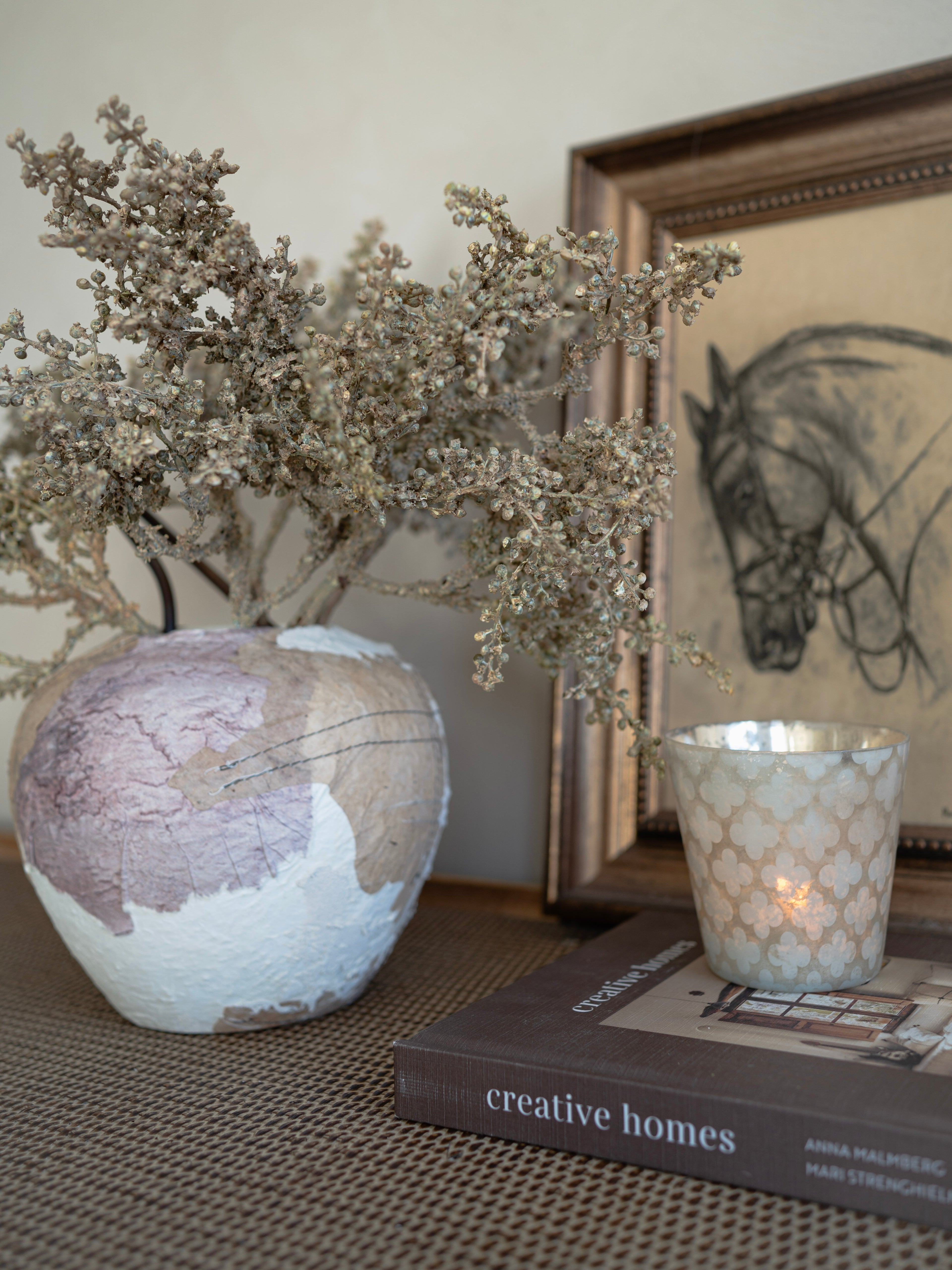 Decorative vase with dried flowers on a surface with a book and candle in the background.