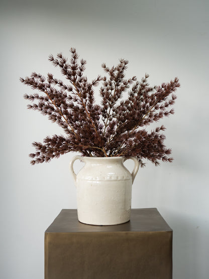 A rustic white ceramic two-handled jug vase filled with dried burgundy-brown ornamental grass, displayed on a dark brown pedestal against a light gray background.