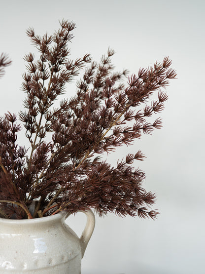 Dried brown ornamental grass stems arranged in a speckled off-white ceramic pitcher against a plain light gray background.