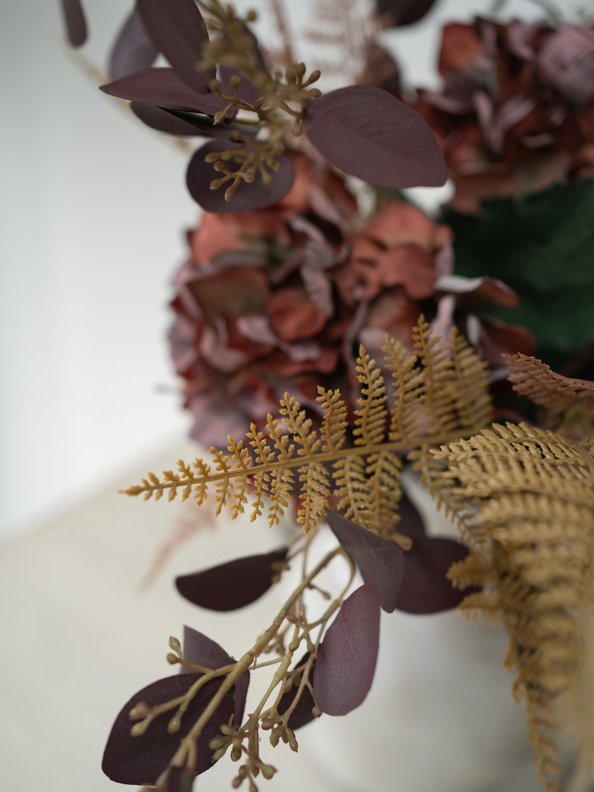 Close-up of a dried floral arrangement featuring muted burgundy hydrangea blooms, dark burgundy eucalyptus leaves, and golden-brown ferns in a white container against a soft white background.