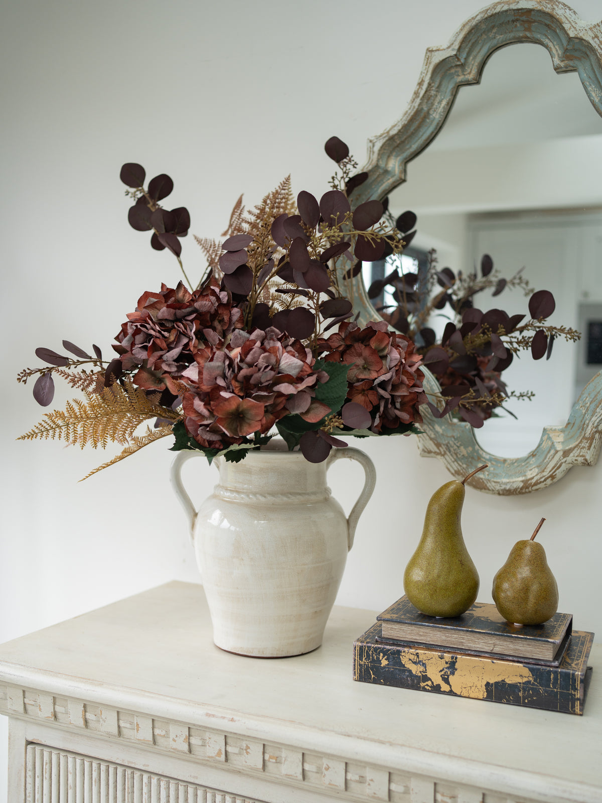 A decorative faux autumn floral arrangement in a white ceramic vase, styled on a white console table next to two green pears and books, with an ornate mirror in the background.