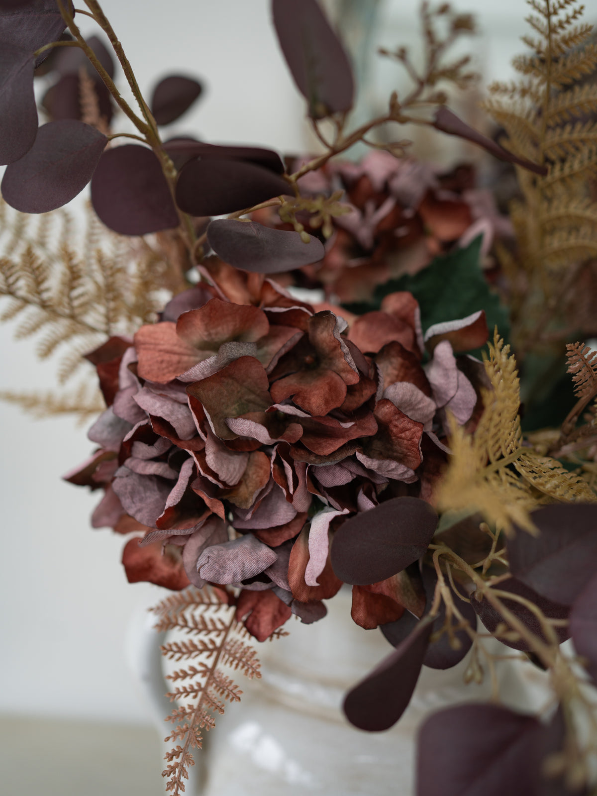 A close-up of a dried faux autumn floral arrangement featuring dusty rose and burgundy hydrangea-like blooms, dark plum eucalyptus leaves, and tan ferns in a white vase.