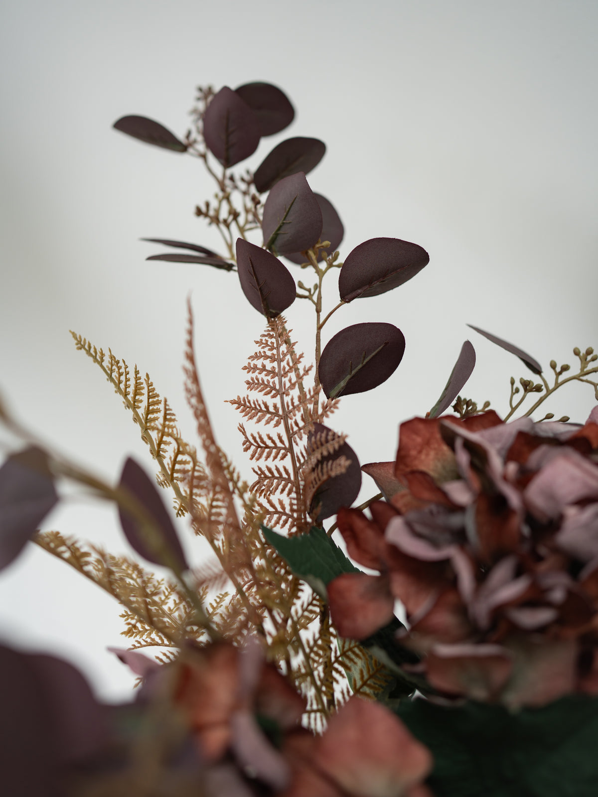 A close-up of faux autumn floral elements including dark burgundy eucalyptus leaves, tan ferns, and mauve-brown hydrangea blooms against a soft light gray background.