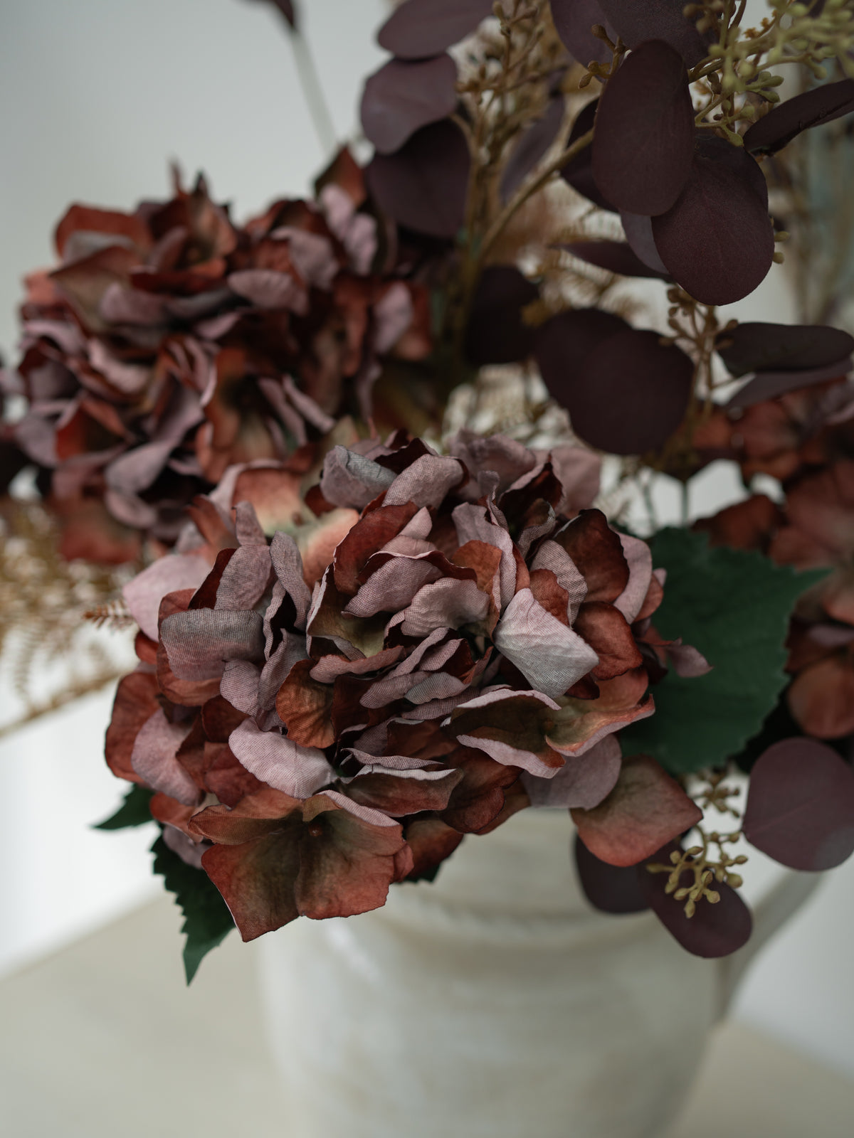 A close-up of a dried floral arrangement featuring large dusty burgundy hydrangea blooms, dark burgundy eucalyptus leaves, and green leaves in a white ceramic pot.
