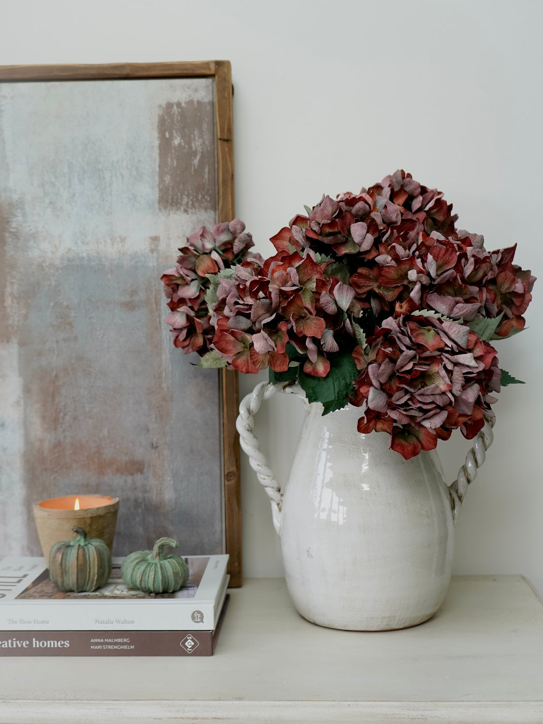 White pitcher with faux brown hydrangeas flowers on a shelf next to a framed picture and books.