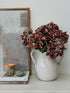 White pitcher with faux brown hydrangeas flowers on a shelf next to a framed picture and books.