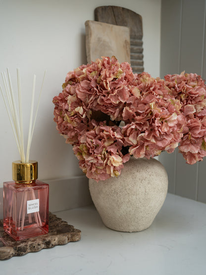 Pink dried effect ruffle hydrangea floral arrangement in a textured vase next to a pink diffuser bottle on a white surface.