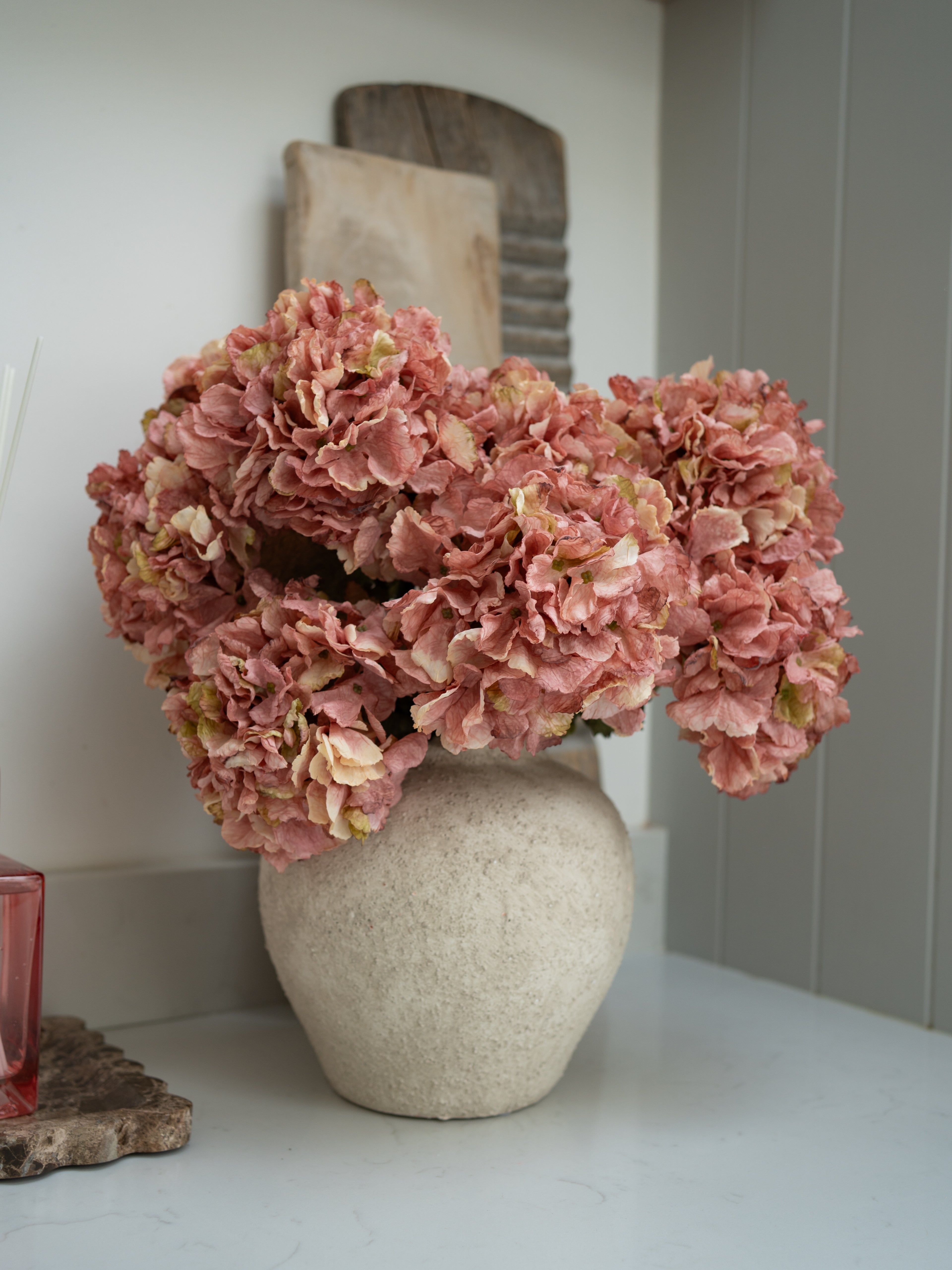 Pink faux dried ruffle hydrangeas in a beige vase on a white surface with a neutral background