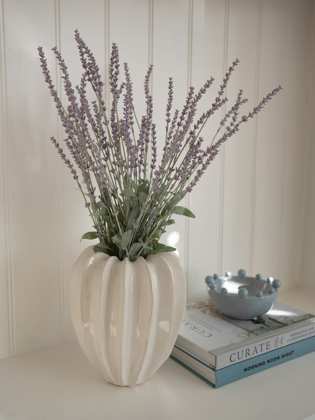 White ridged ceramic vase with faux lavender bush on a surface with books and a small pale blue bobble edged bowl.