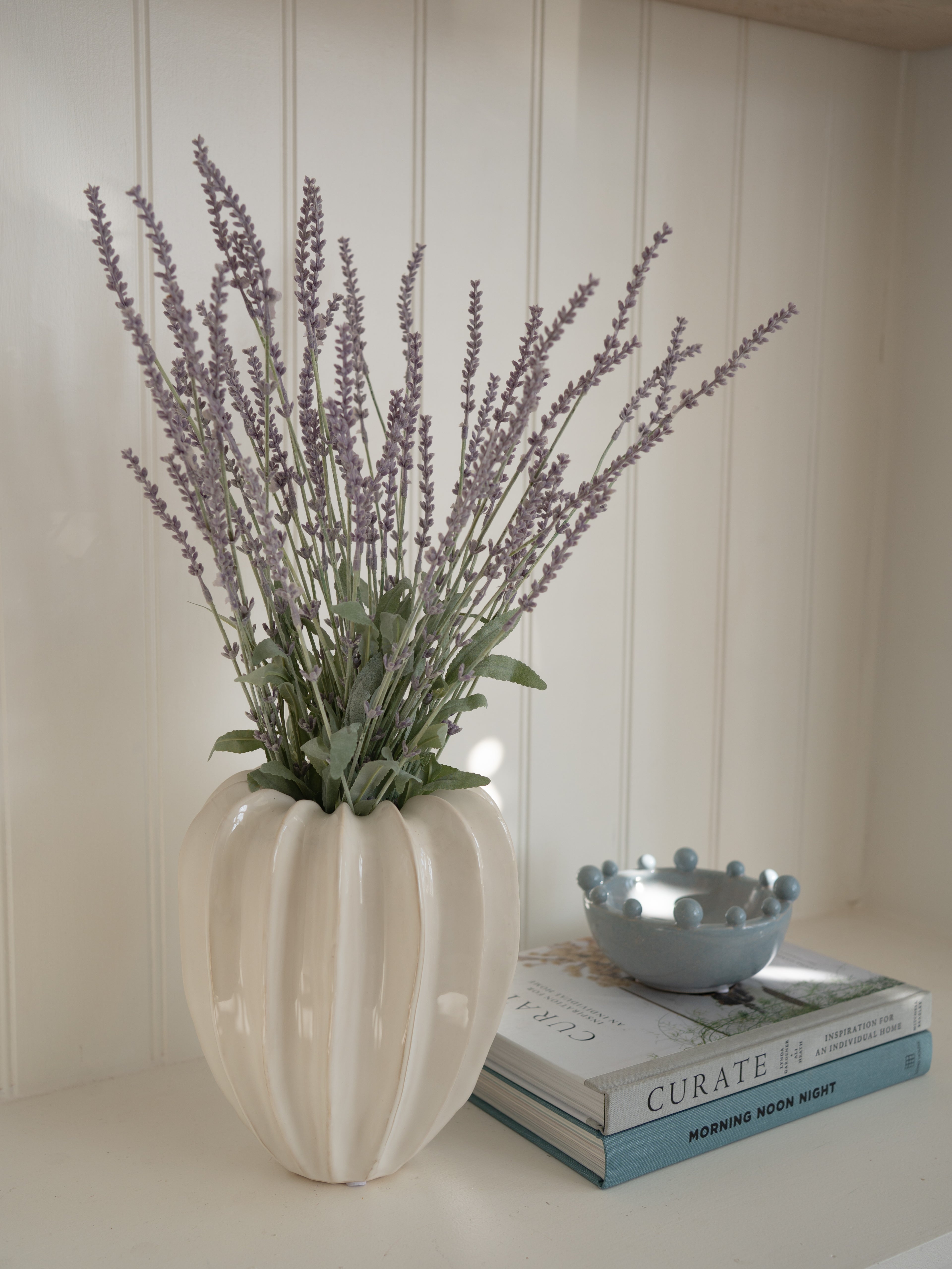 Decorative vase with faux lavender on a surface with books and a bowl.