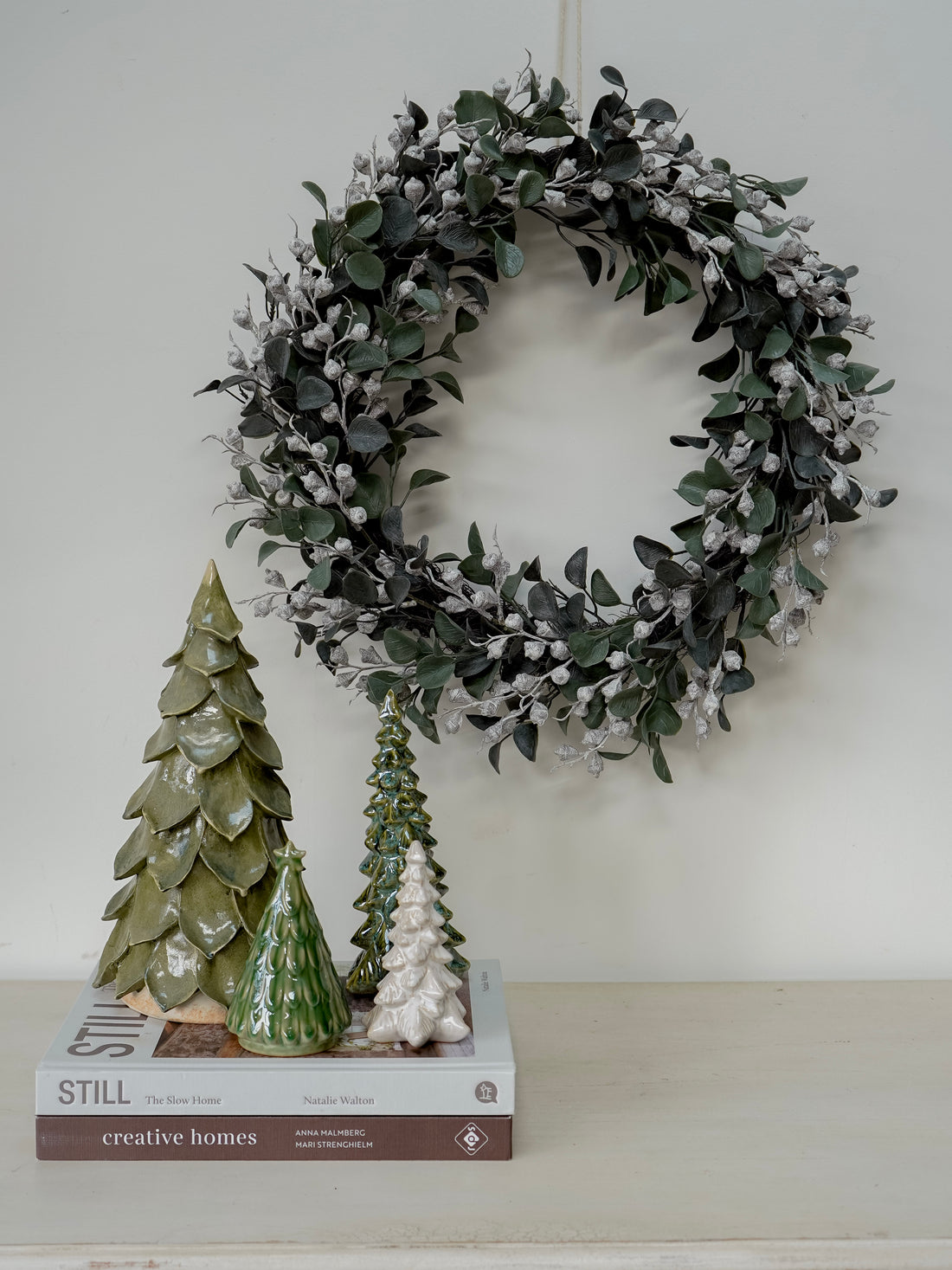 Decorative wreath with faux christmas greenery and small trees on a shelf against a white wall.