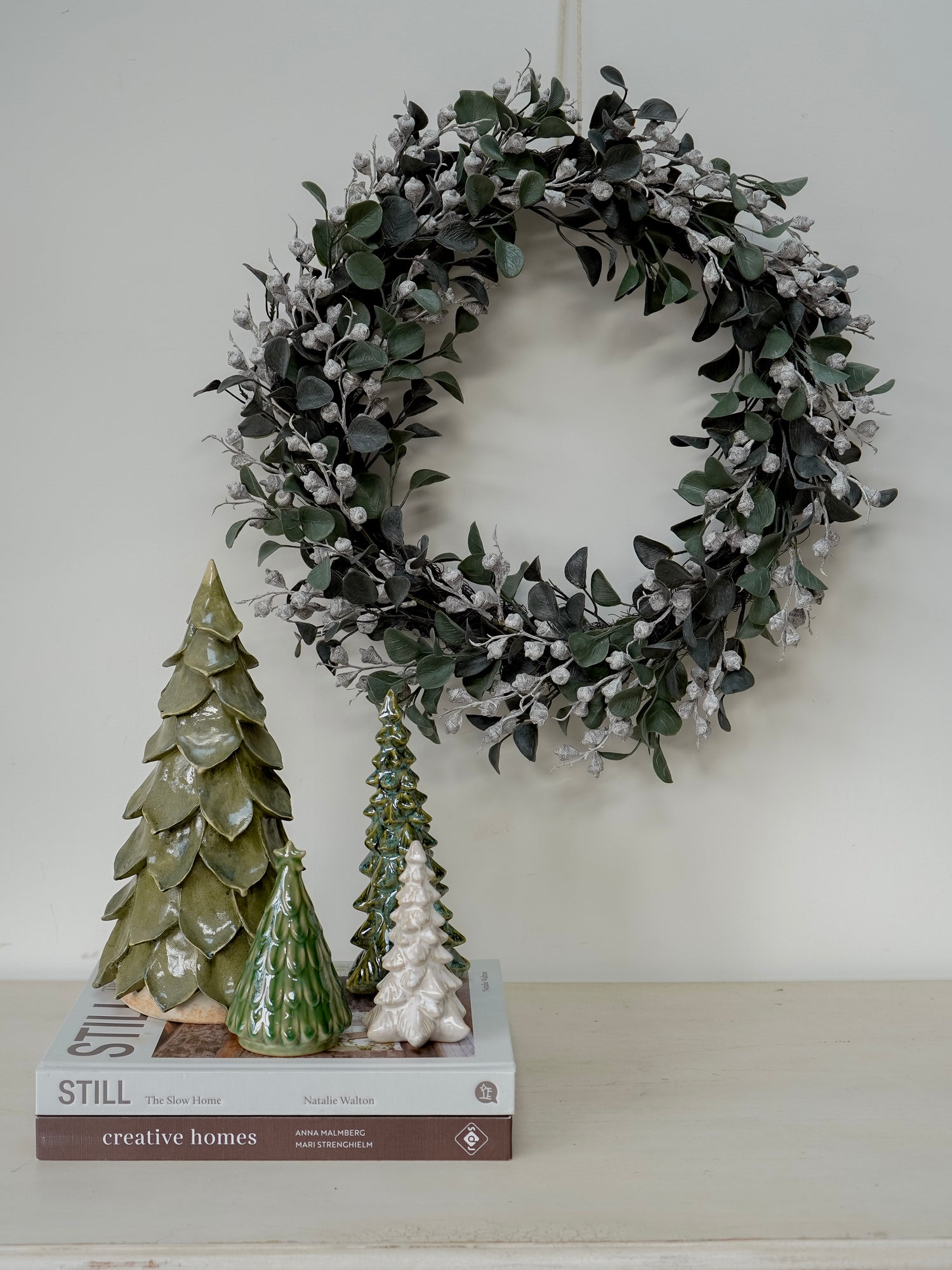 Decorative wreath with faux christmas greenery and small trees on a shelf against a white wall.