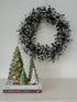 Decorative wreath with faux christmas greenery and small trees on a shelf against a white wall.