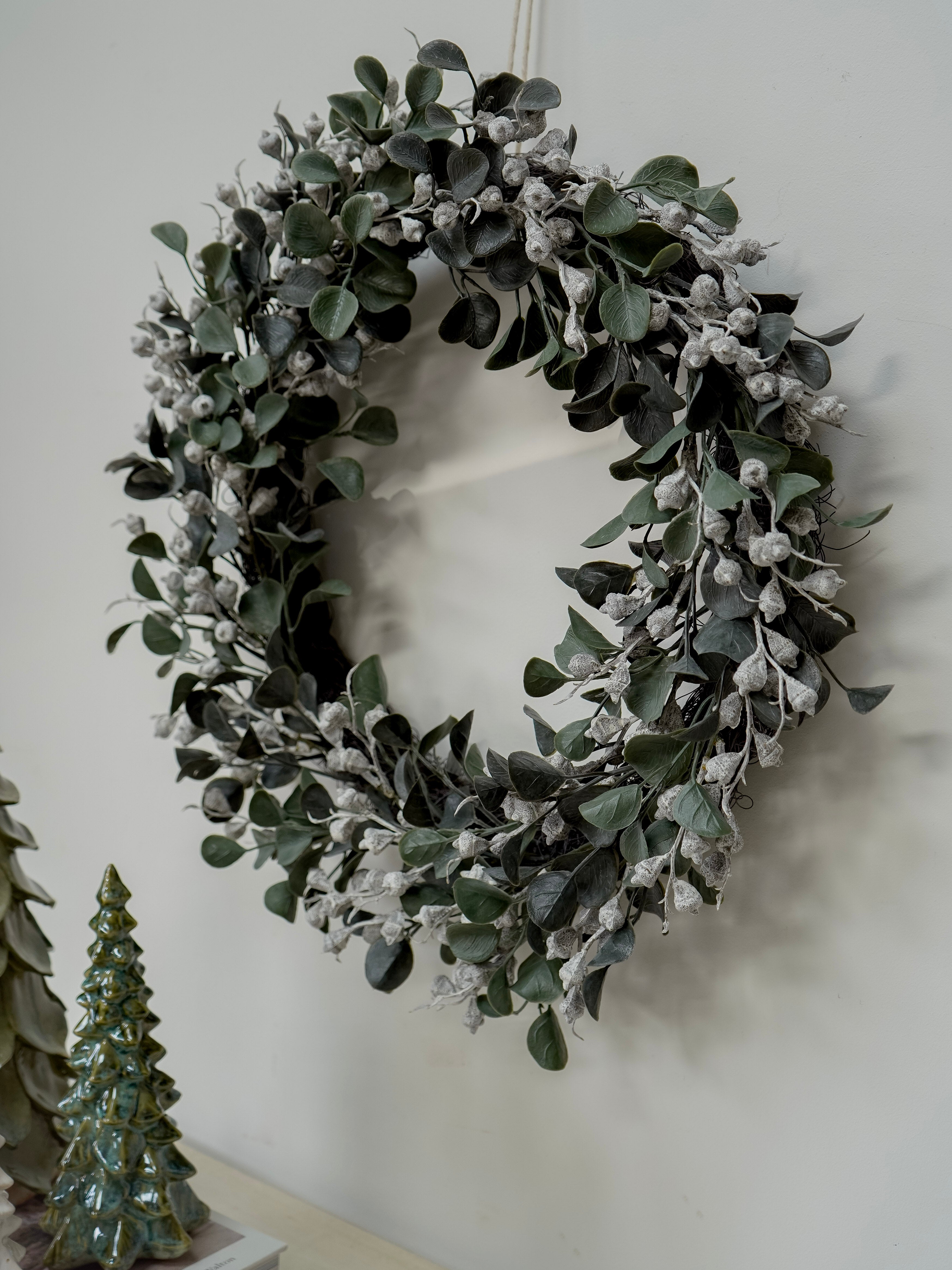 Green leafy wreath on a white wall with a small decorative tree in the corner.