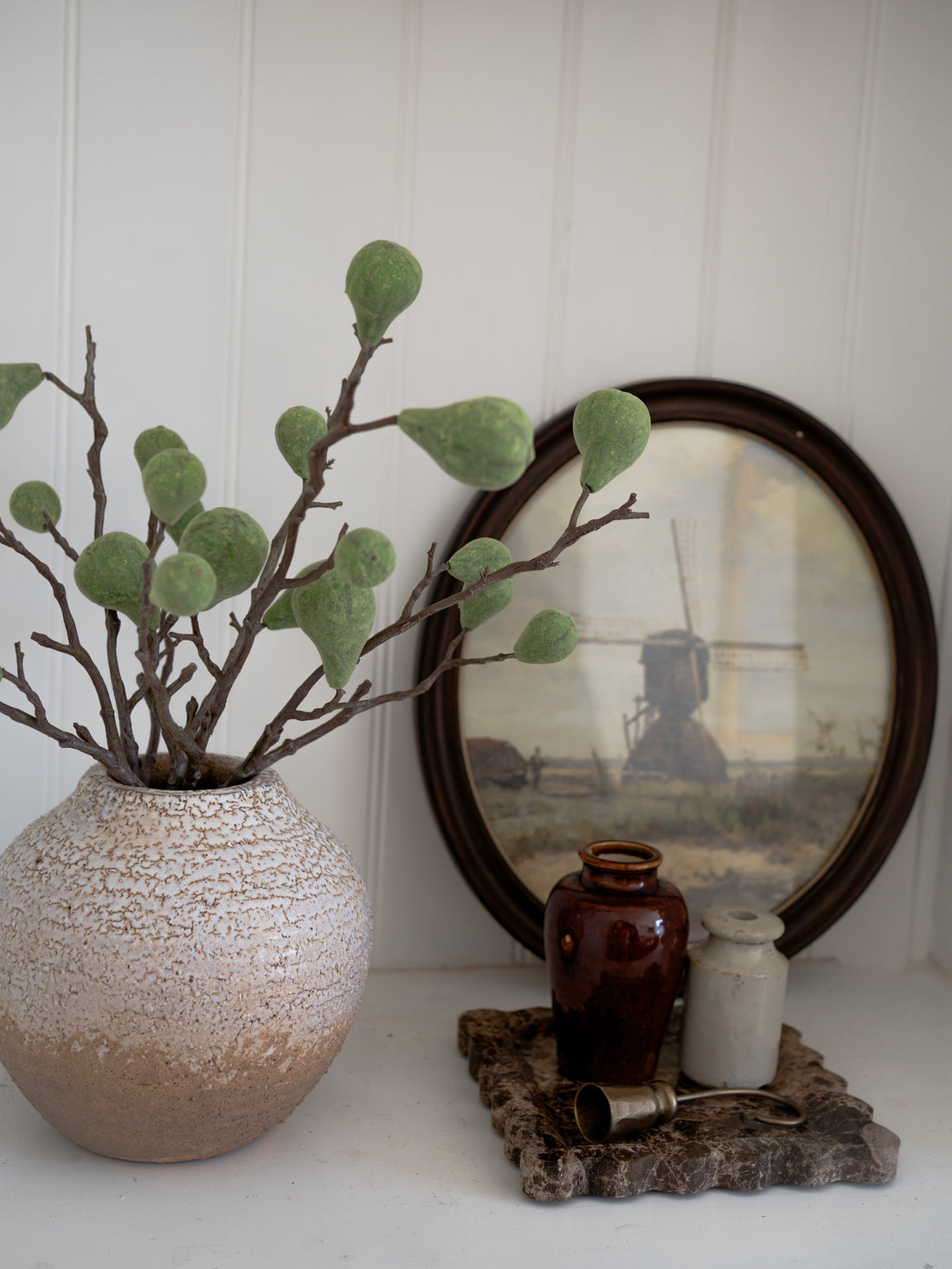 Decorative setting with a textured vase, faux green flocked fig branches, and small bottles on a white surface.