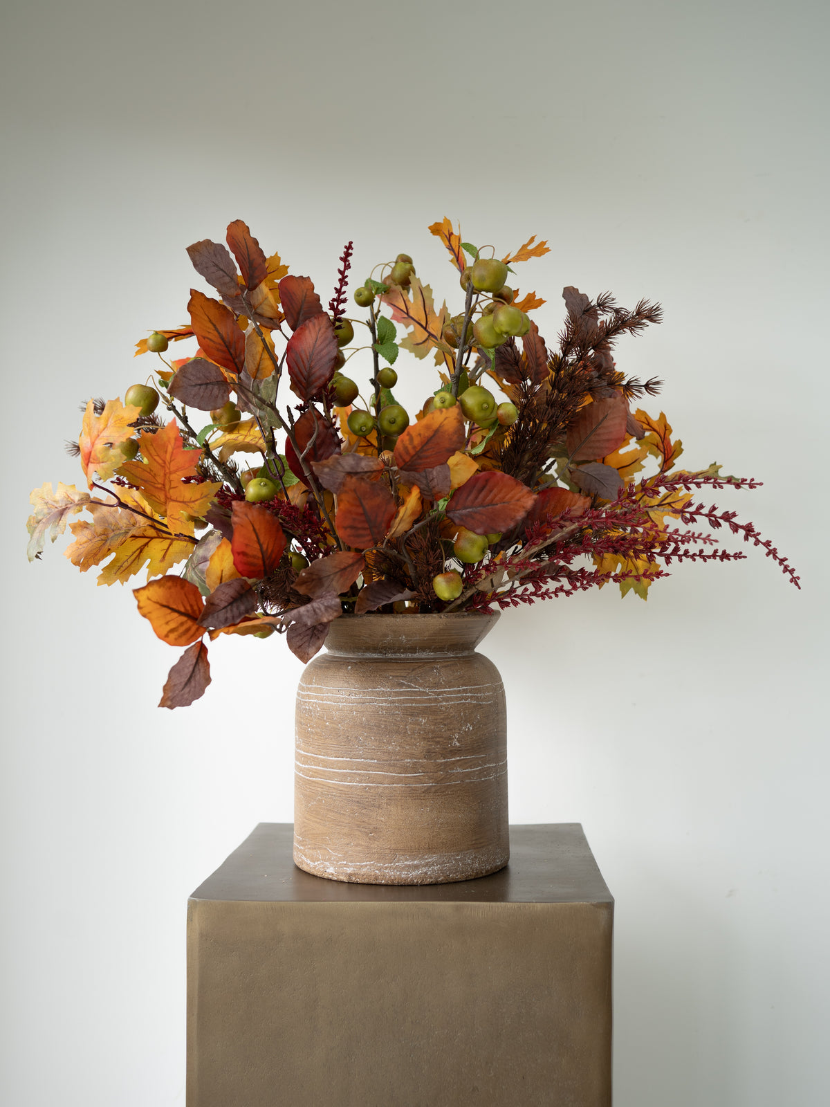 An autumn floral arrangement with orange, brown, and yellow leaves, small green berries, and burgundy stems, displayed in a rustic beige ceramic vase on a square pedestal against a light gray background.