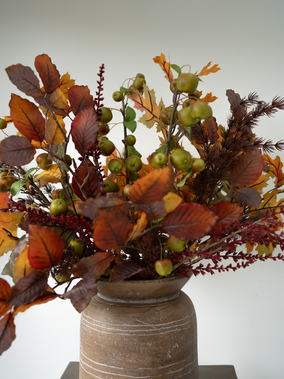 An autumn floral arrangement featuring faux leaves in shades of orange, brown, and red, small green apples, and dark brown pinecones, presented in a rustic brown ceramic vase against a plain light gray background.