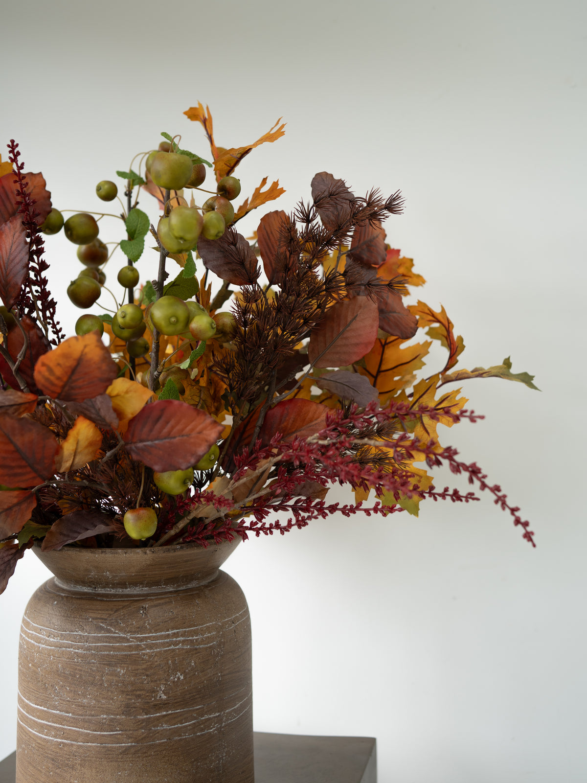 An autumn floral arrangement featuring orange and brown leaves, green berries, and burgundy stems, displayed in a rustic brown ceramic vase against a plain light gray background.