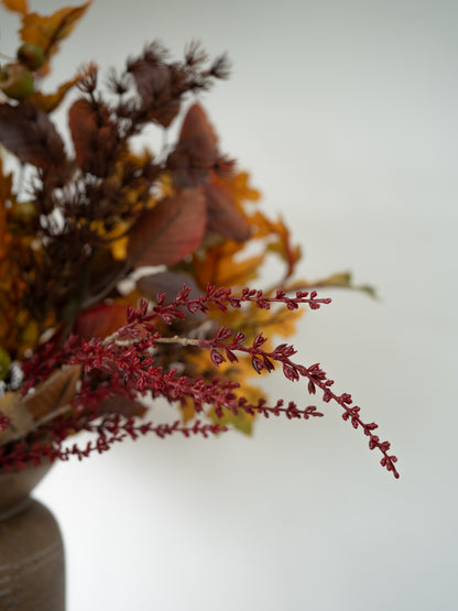 A close-up of dried autumnal floral stems with burgundy-red berries and warm brown, orange, and yellow leaves, arranged in a neutral-toned vase against a soft light gray background.