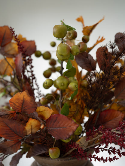 An autumn artificial floral arrangement featuring faux green apples, orange and brown leaves, and burgundy dried stems in a rustic container against a neutral light gray background.