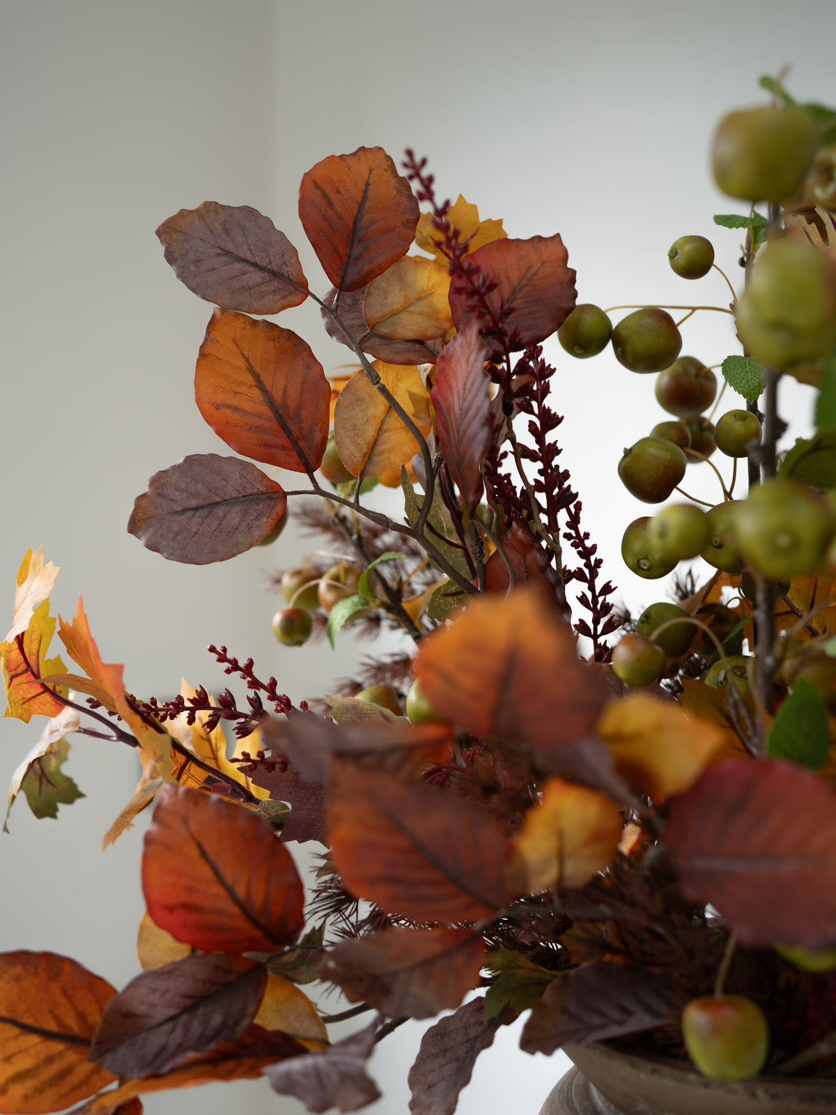 An autumn-themed artificial floral arrangement featuring brown and orange leaves, burgundy berries, and green apple-like fruits, set against a plain light gray background.