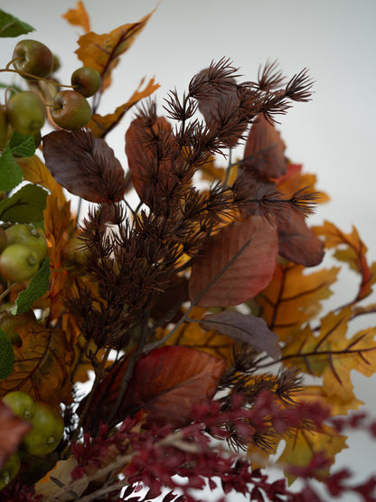 An autumn floral arrangement featuring dark brown pinecones, yellow and orange maple leaves, green apples, and burgundy berries against a soft, light gray background.