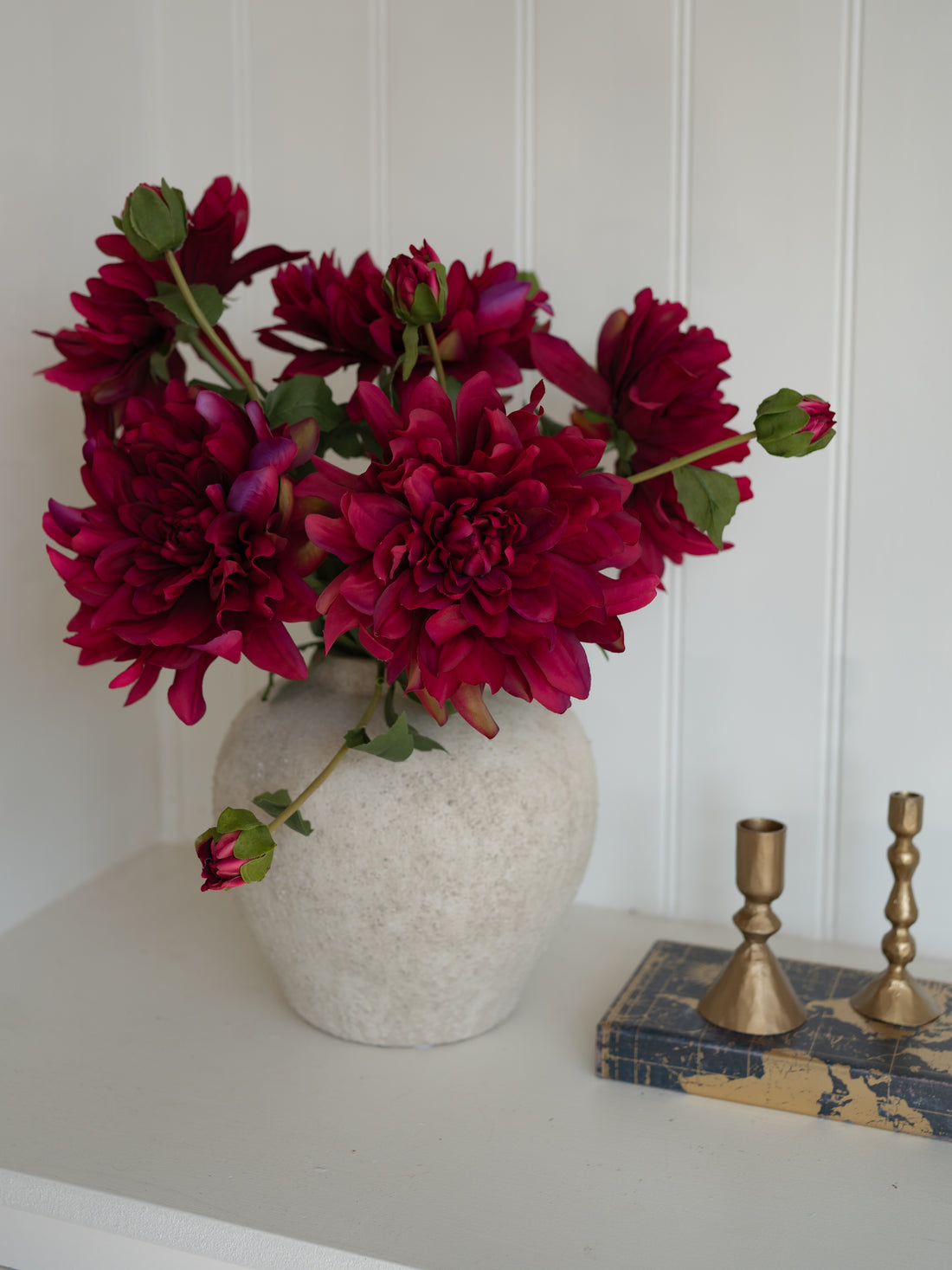 Vase with Faux Fuchsia Dahlia flowers on a white surface with gold candlesticks in the background