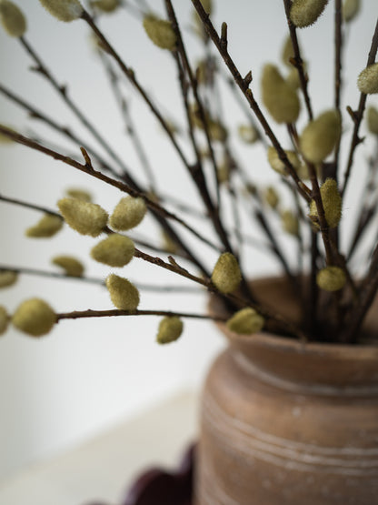 Artificial willow branches with fuzzy light green buds arranged in a rustic brown ceramic pot against a soft white background.